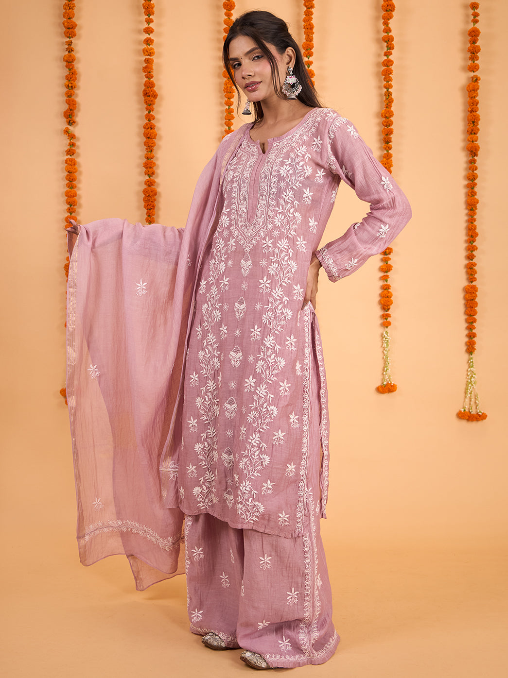 Model showcasing a Mul Chanderi Chikankari kurta set with decorative neckline borders, embroidered sleeve cuffs and flowing palazzo pants against a marigold backdrop.