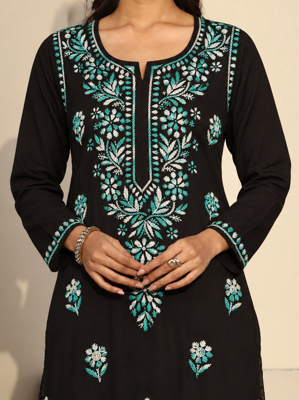 Model wearing the Ayra Chikankari kurti against a textured wall, showing its straight cut, embroidered sleeve edges, scattered floral motifs and long hem falling below the knees.