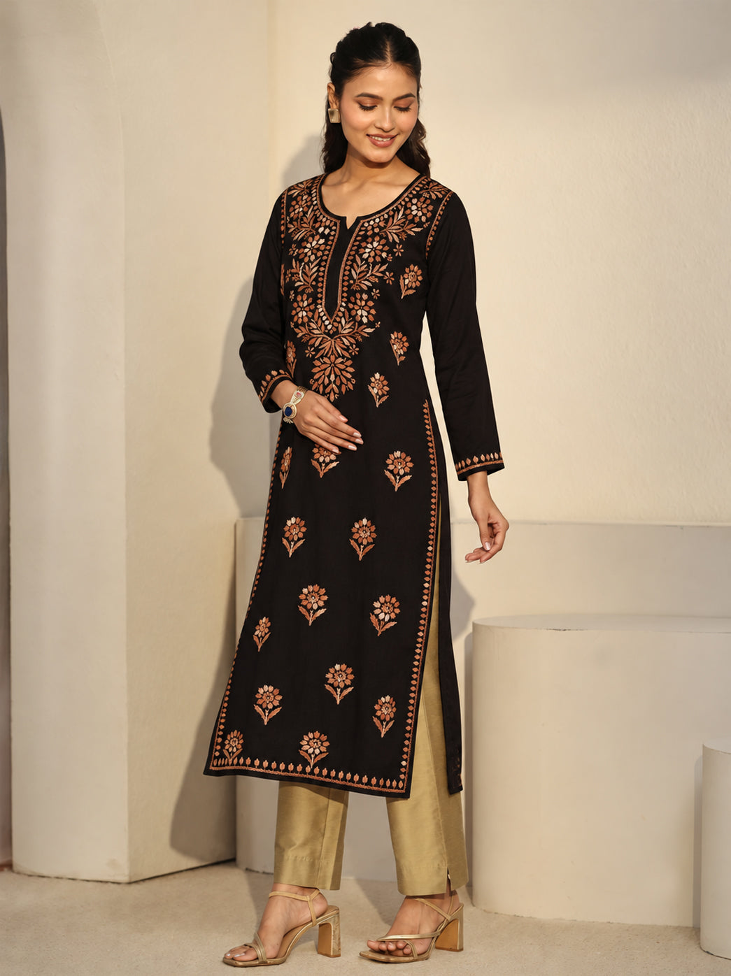 Model wearing the Ayra Chikankari kurti against a textured wall, showing its straight cut, embroidered sleeve edges, scattered floral motifs and long hem falling below the knees.