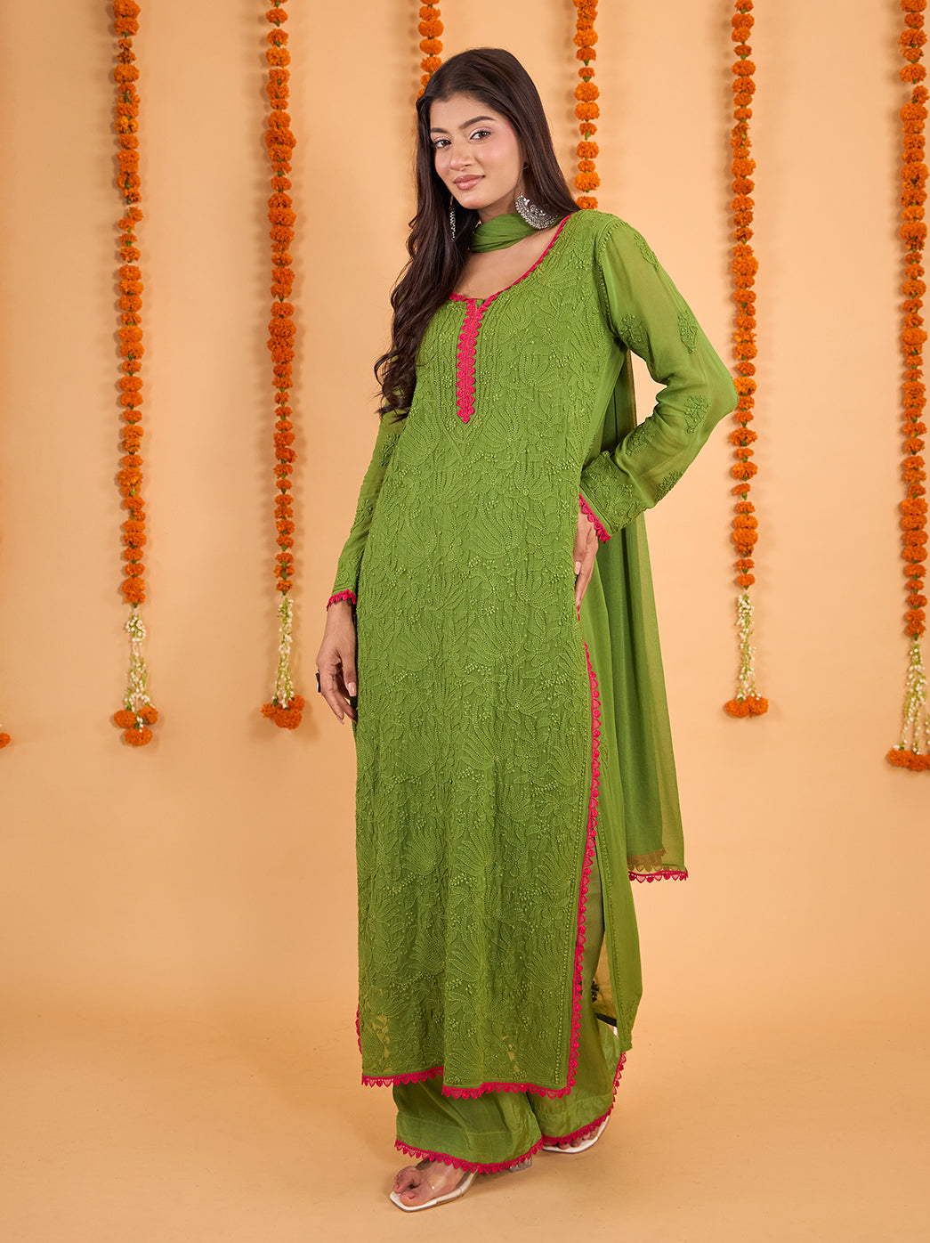 Model showcasing the Gulnaar Chikankari festive wear set against a backdrop of hanging marigold garlands, with the kurti embroidery clearly visible.

