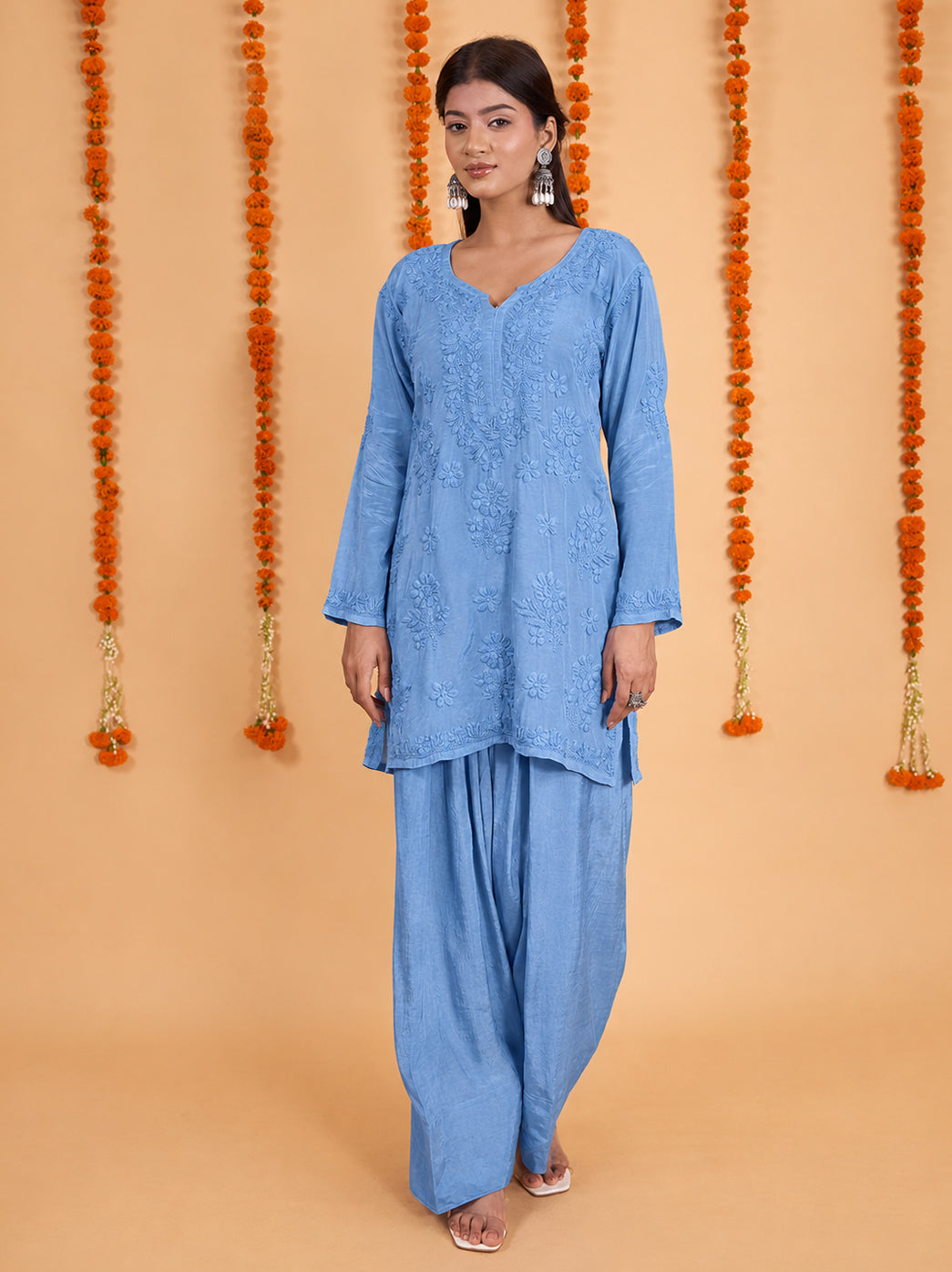 Model wearing the Shagun Muslin Chikankari Kurti Farshi Set, featuring a floral embroidered short kurti and flowing Farshi pants against a backdrop of hanging marigold garlands.