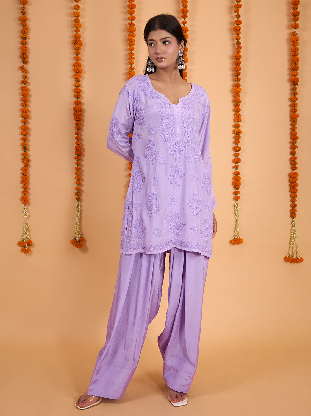Model wearing the Shagun Muslin Short Kurti Farshi Set, featuring floral Chikankari embroidery and flowing wide-leg pants against a marigold-decorated backdrop.
