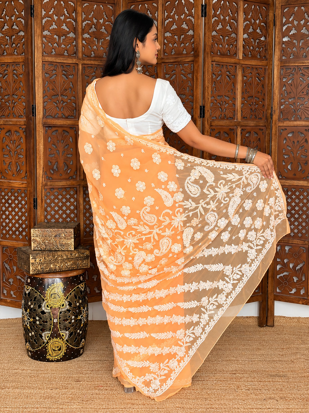 Srija Georgette Chikankari Saree styled with a short-sleeved blouse and statement earrings, with the embroidered pallu displayed against carved wooden panels.